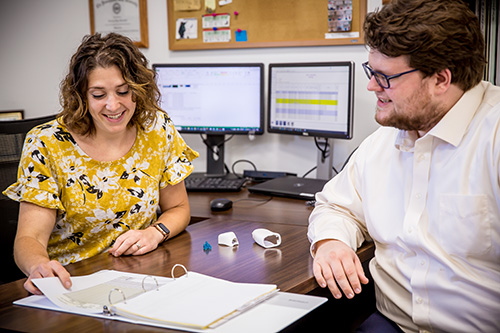 Woman sitting at a desk, talking to a man while she is looking through a book